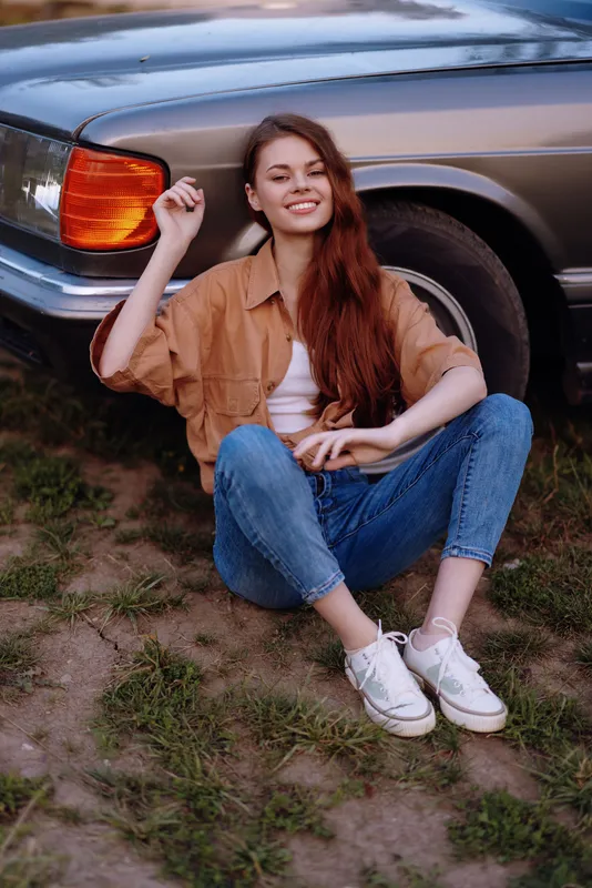 Young smiling woman sitting beside vintage car, wearing casual outfit with a relaxed expression in natural setting with earthy tones.