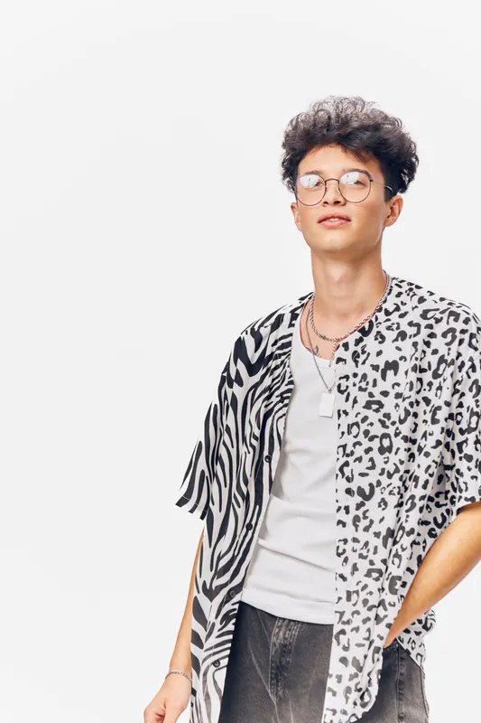 young man wearing a black and white patterned shirt with glasses and curly hair, standing confidently against a bright white background