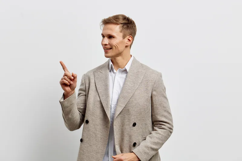 Confident young man in stylish beige blazer and white shirt pointing with finger while smiling, standing against plain white background. Studio portrait, fashion, success, communication, professional,