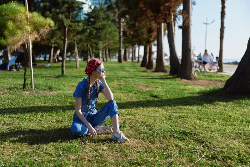 young woman sitting on grass in park wearing blue outfit and red flower headband enjoying sunny day with trees in background
