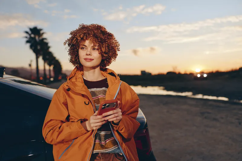 Woman holding smartphone outdoors at beach sunset, curly hair, warm orange jacket, relaxed smile, lifestyle moment during golden hour, soft ocean breeze, scenic coastline background
