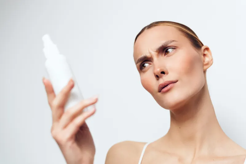 Young woman with clear skin holding white cosmetic bottle close to face, examining product carefully, neutral background, natural lighting, skincare concept, focused expression.