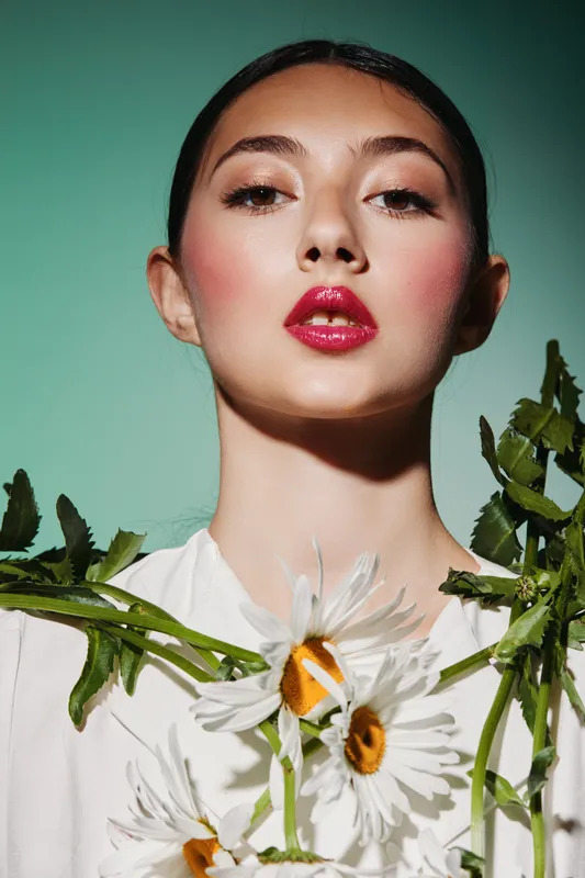 Portrait of a fashionable young woman with glossy red lips and glowing skin wearing white dress decorated with large daisies. Studio shot on a green background. Creative beauty concept.