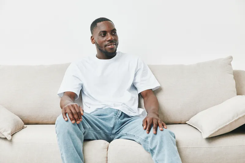 A young Black man sitting comfortably on a beige sofa, wearing a white t shirt and jeans, exuding a relaxed and cheerful emotion in a cozy living room setting