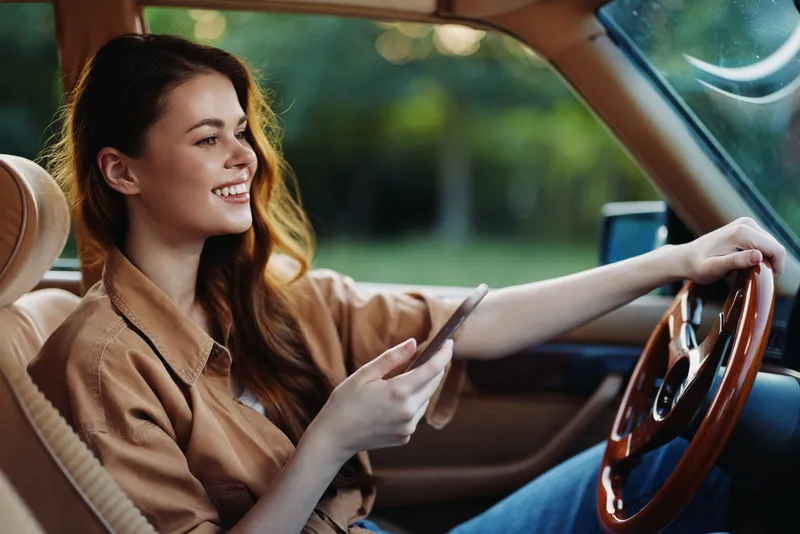 Cheerful young woman enjoying a smartphone while driving in a vintage car, showcasing a relaxed atmosphere with green nature background, bright colors, and casual fashion.