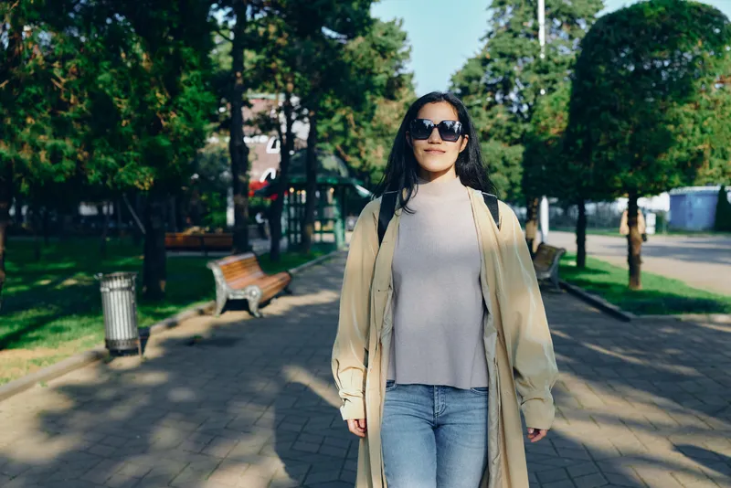 Woman walking in a sunny park wearing a beige coat and light sweater, sunglasses, jeans, and confident stride, outdoor leisure and fashion moment on a tranquil path