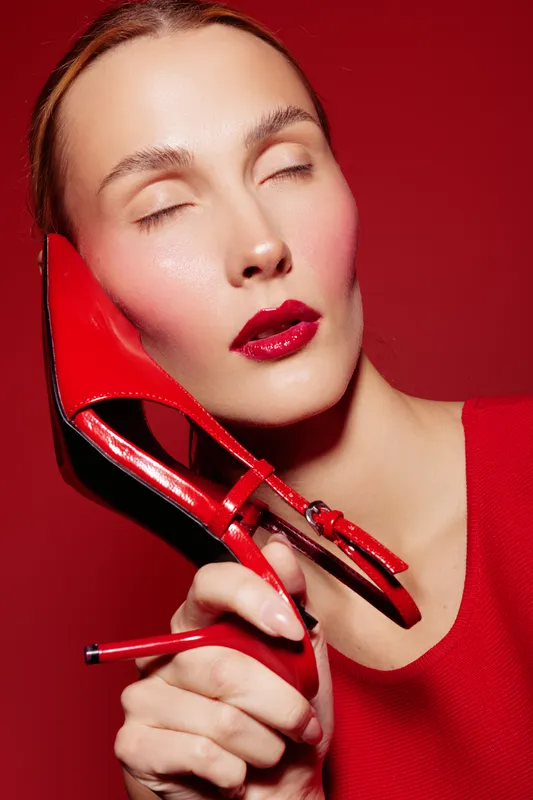 Close-up of a woman with closed eyes holding a red high heel shoe near her face against a red background. Concept of fashion, beauty, and bold makeup with glossy red lips.