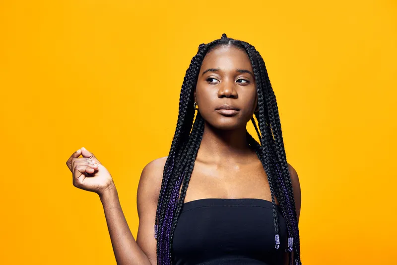 A confident young woman with braided hair poses against a vibrant yellow background, showcasing her unique style and expressive emotions, perfect for lifestyle themes