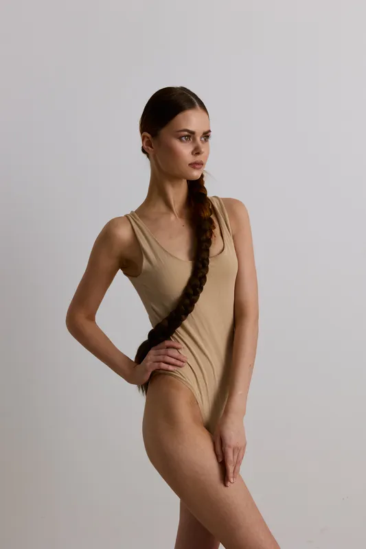 Slender woman with long braided hair wearing a beige bodysuit, posing gracefully against a plain light background, highlighting natural beauty and elegance.