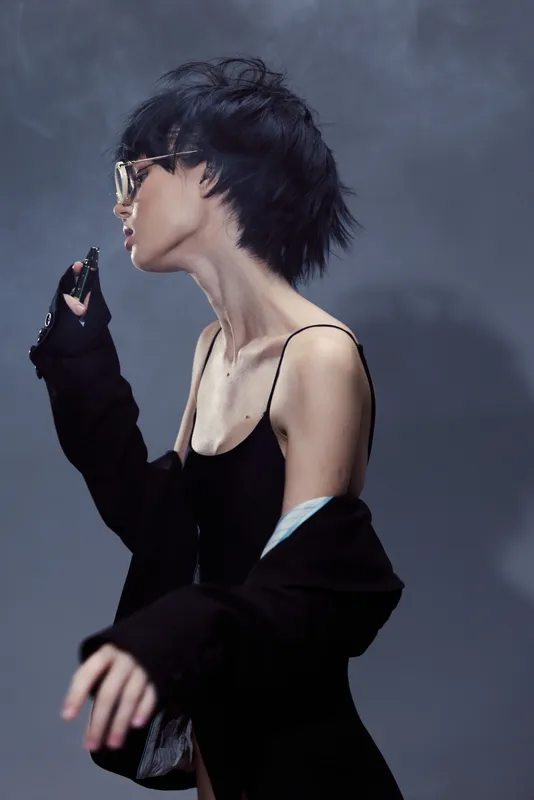 Stylish woman with short dark hair wearing glasses and black clothing in a minimalist moody setting.