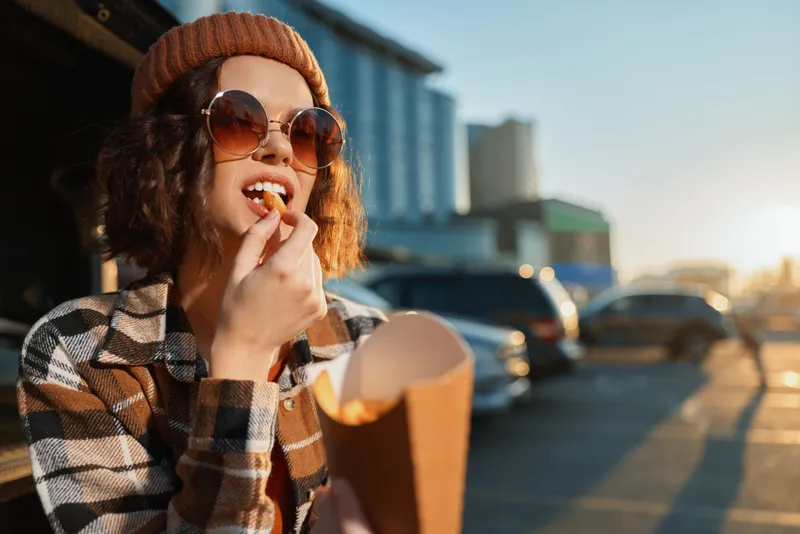 woman fries street sunglasses beanie sunlight candid urban - Young woman in beanie and sunglasses eating fries on a city street at golden hour, authenticity, candid lifestyle, golden hour glow,