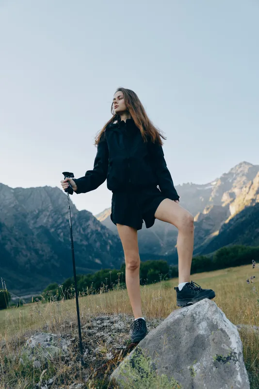 A hiker stands atop a rock with a trekking pole, gazing toward distant mountains under clear skies, embodying adventure, resilience, and outdoor spirit in a rugged landscape.