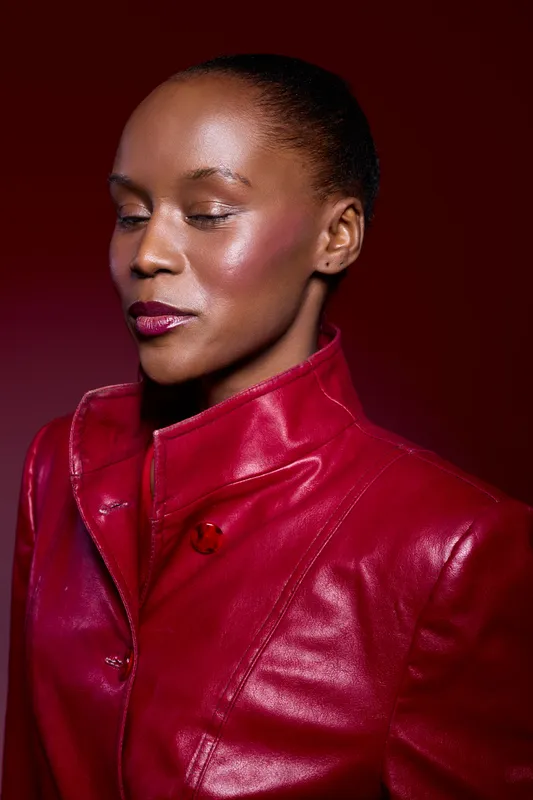 Portrait of confident African woman with dark skin wearing red leather jacket on dark red background. Studio lighting highlights natural skin texture and calm facial expression.