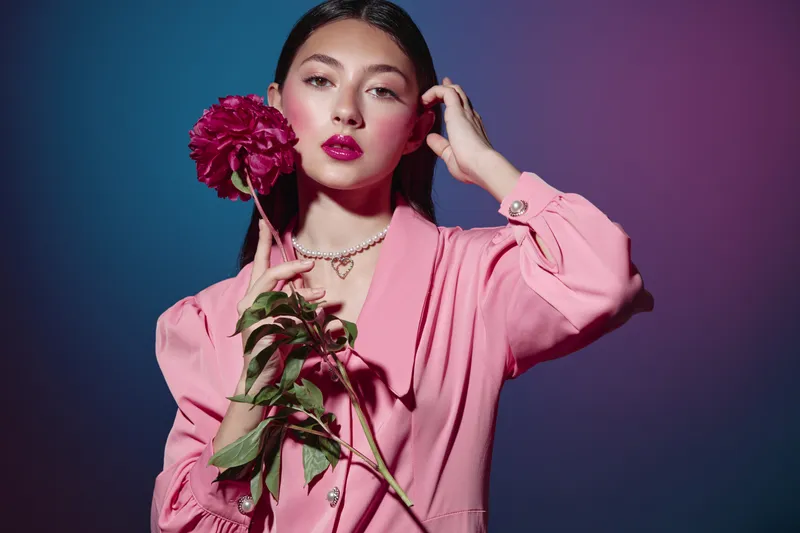 Beauty model wears pink blazer, holding flower close to cheek. Glamorous makeup and elegant pose with soft purple and blue background. Creative fashion studio shot.