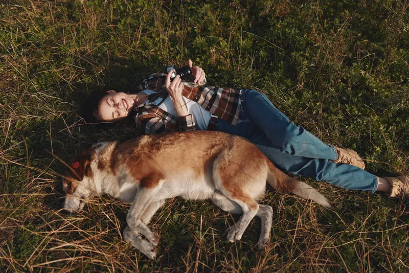 Woman lying on grass with a friendly dog, relaxing outdoors in a sunny park, casual attire, nature scene, gentle breeze, slow moment, leisure and companionship