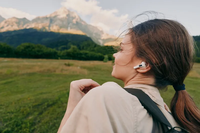 Woman enjoying outdoor landscape with headphones, hiking past mountains, casual backpack, serene mood, bright natural light, lifestyle moment in open field and meadow