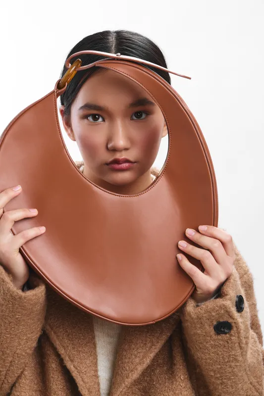 asian woman wearing a warm brown coat holds a circular leather bag around her face, creating a bold fashion portrait with a minimalist studio backdrop.