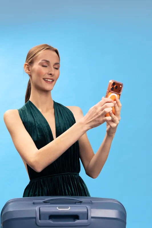 Young woman in dark dress smiling and holding smartphone taking selfie near suitcase on blue background. Concept of travel, vacation, tourism, and modern lifestyle with happy mood.