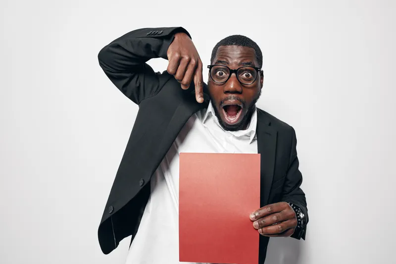 African American man with dark skin and glasses in black suit pointing down at red folder with surprised expression on white background. Business, office and communication concept.