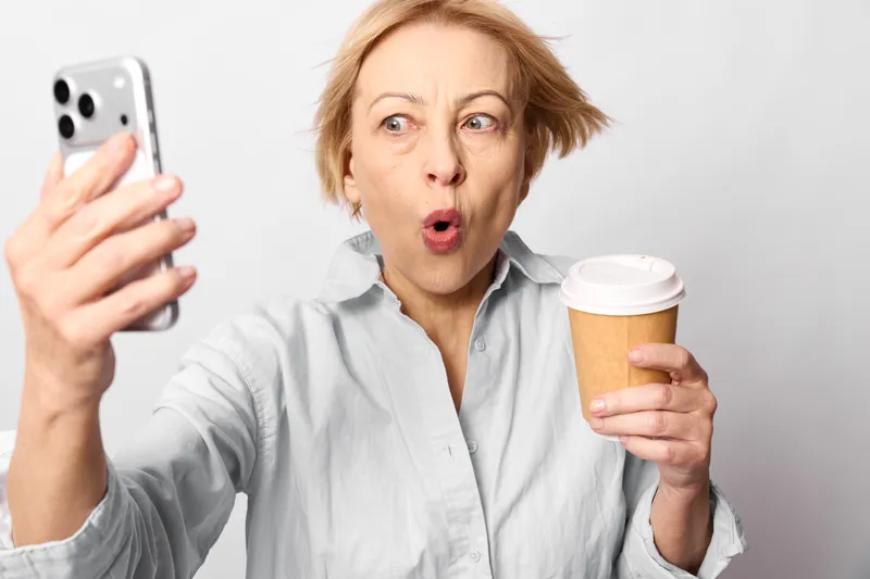 Surprised woman holding smartphone and coffee cup, wide open mouth expression, looking at screen, white background, casual home scene, morning caffeine moment