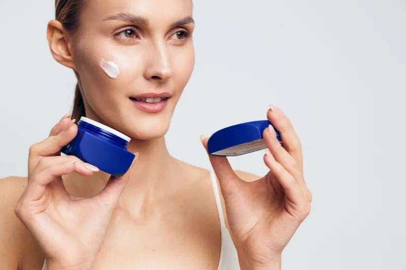 Woman holding blue moisturizer jar and cosmetic lid, applying face cream to cheek, calm expression, clean studio background, close up beauty portrait, fresh healthy skin