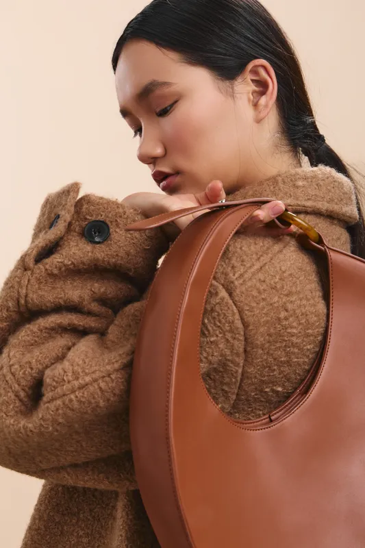 asian woman wearing a cozy coat embraces a plush bear while holding a brown leather bag in a soft studio setting, conveying warmth and gentle comfort.