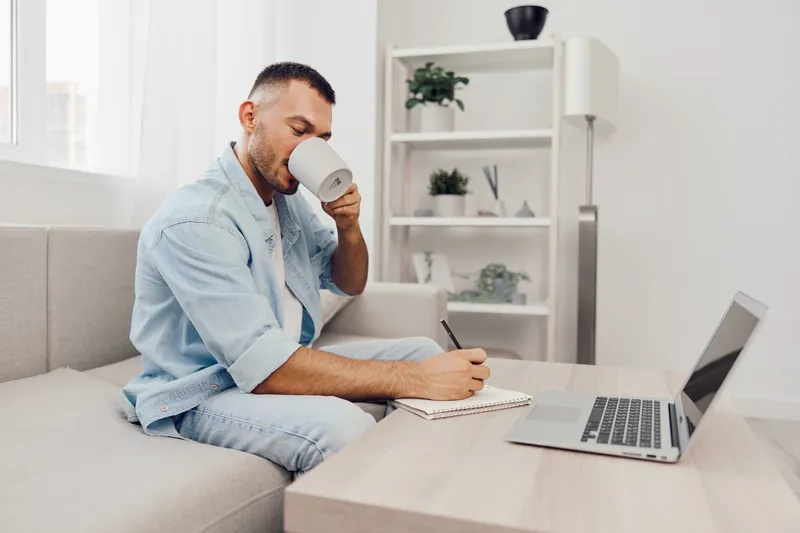 A focused man enjoying coffee while writing in a notebook, sitting on a cozy couch in a modern living room. The atmosphere inspires productivity and calmness.