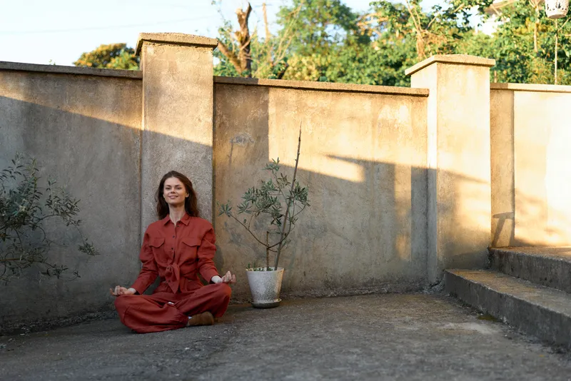 Woman lifestyle scene, seated cross legged in a red outfit on a sunny patio, serene mood, simple backdrop, casual pose, peaceful outdoor moment, bright day lighting
