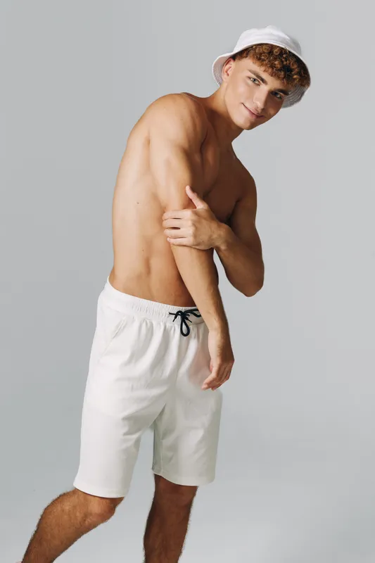 Young man with curly hair wearing white shorts and bucket hat posing shirtless on gray background with confident expression and casual summer style.