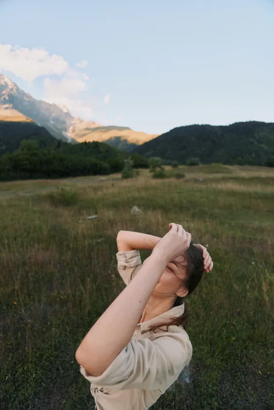 Young woman in a light jacket stands in a grassy field with distant mountains, arms raised, enjoying fresh air and warm sunlight in nature.