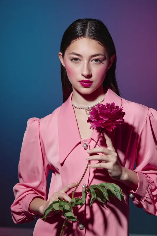 Young woman in pink satin blouse holding dark pink flower. Fashion-inspired beauty portrait with dramatic colorful lighting and elegant pose. Concept for creative fashion and beauty art.