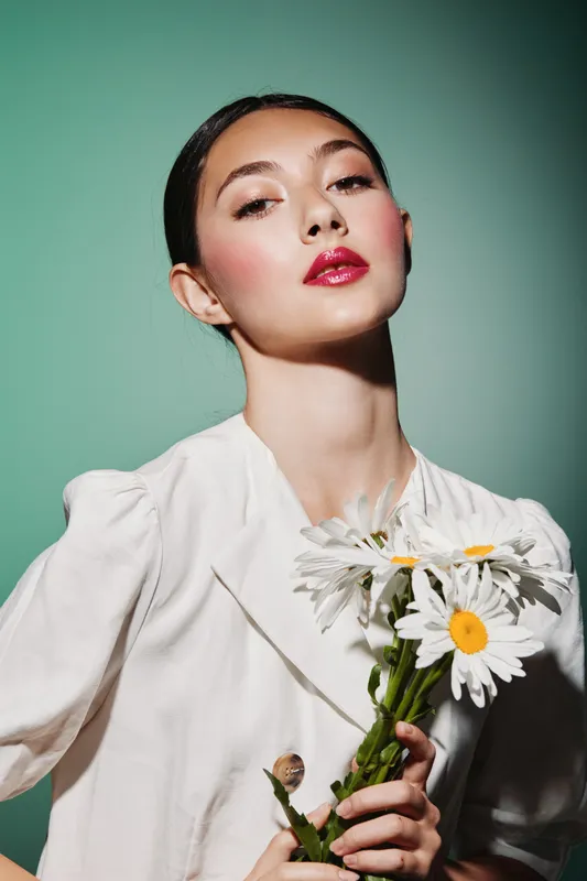 Elegant young woman with flawless makeup and glossy crimson lips poses confidently holding a bouquet of white daisies. Fashion studio lighting emphasizes her smooth skin beauty creative concept.