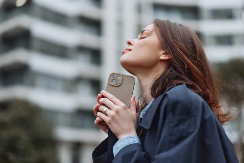 Young woman with long brunette hair enjoys a peaceful moment outdoors holding a smartphone close to her chest with closed eyes and relaxed expression.