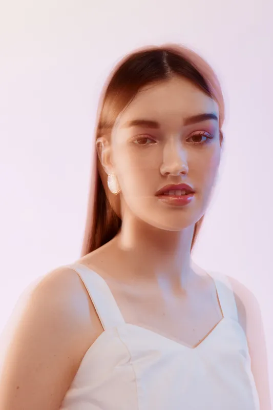 Young woman with long hair wearing white top poses in soft lighting. Beauty and fashion concept with delicate makeup and calm expression on pastel background.