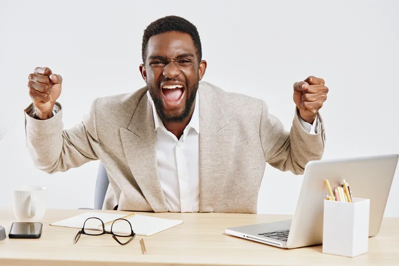Confident excited young African American man in beige blazer and white shirt celebrating success sitting at modern desk with laptop and glasses isolated on plain white background.