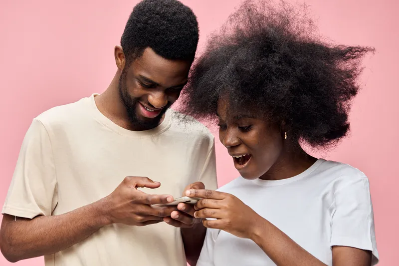 Happy young couple interacting with a smartphone on a pink background, showcasing joy and connection with modern technology, wearing casual attire