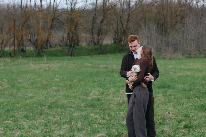 Young couple embraces outdoors while holding a small white baby goat in a grassy field during autumn, expressing warmth and connection in a natural lifestyle setting.