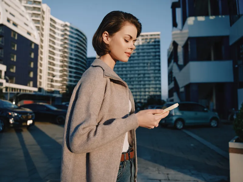 A stylish woman stands outdoors in an urban setting, focused on her smartphone. She wears a neutral coat and casual attire, capturing a modern, on-the-go moment with calm confidence.