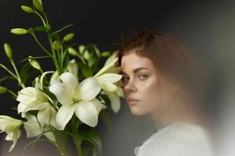 A young woman with brown hair gazes pensively at the camera while holding white lilies, reflecting a serene and natural aesthetic adorned with soft colors