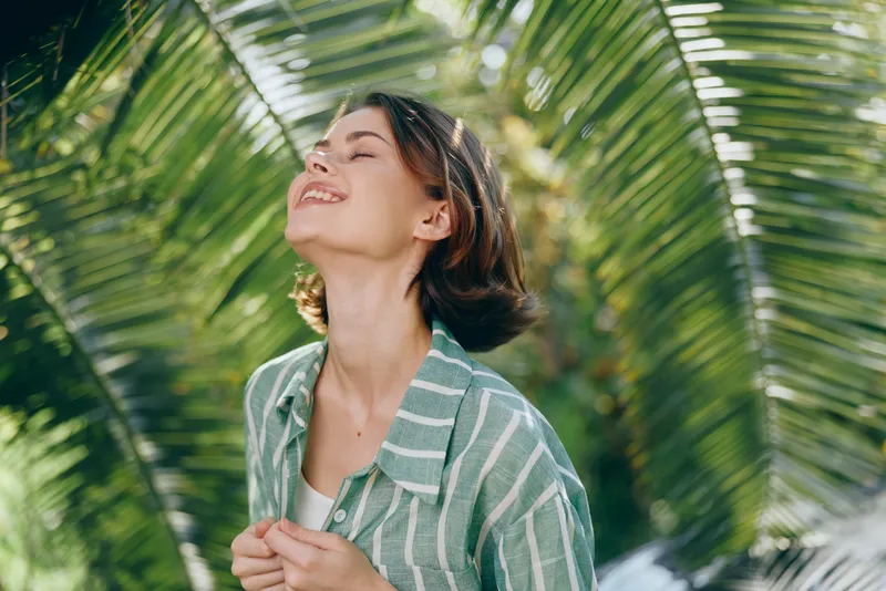 A joyful woman stands outdoors with a bright smile, enjoying warm sunlight and a gentle breeze.