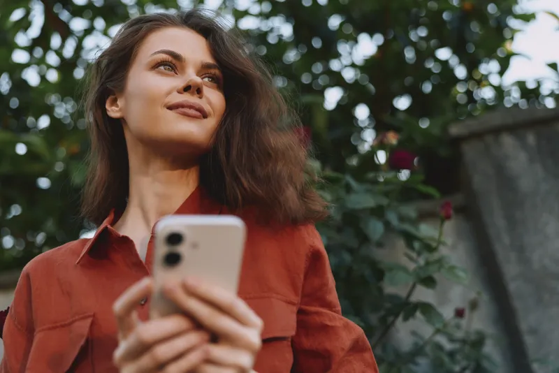 Woman with brown hair in a rust shirt holding a smartphone outdoors. Close-up on greenery and soft natural light, conveying modern lifestyle and confidence everyday