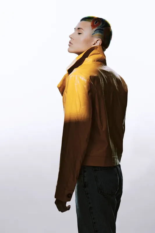 Woman with colorful shaved hair wearing a yellow jacket and denim jeans, standing in profile with eyes closed against a light background, stylish urban fashion.