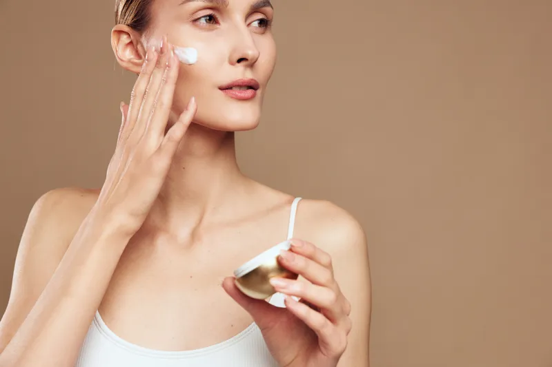 Young woman with clear skin applies face cream on cheek using fingers holds cosmetic jar isolated on brown background skincare beauty routine concept closeup portrait.