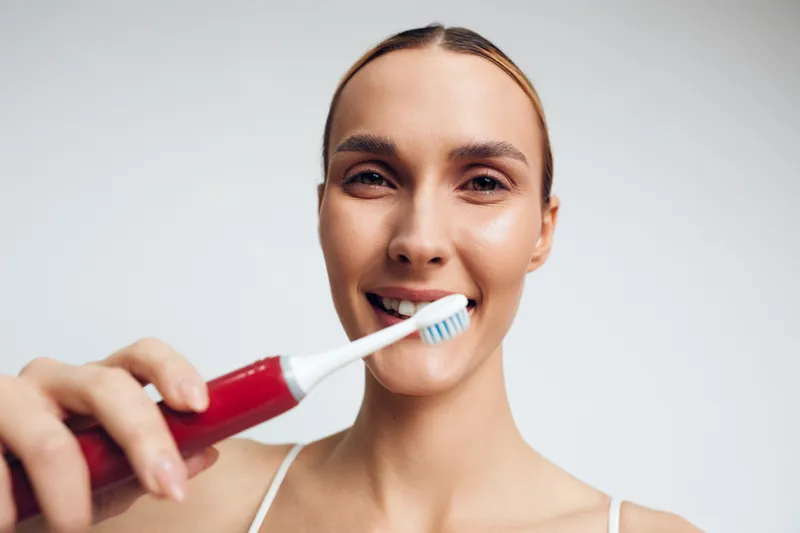 Young woman brushing teeth with electric toothbrush indoors, close up portrait, natural smile, clean skin, white background, morning hygiene routine