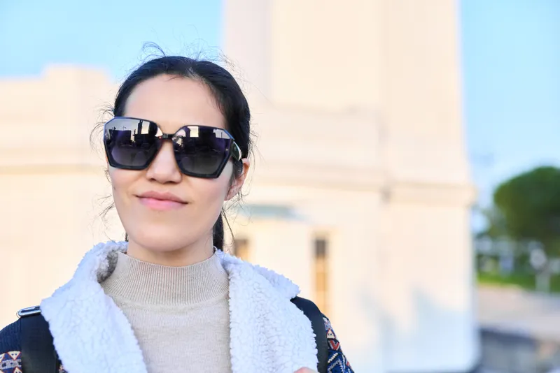 Sunglasses, woman, outdoor fashion scene featuring light scarf and sweater, warm sunlight, urban backdrop, relaxed expression and confident pose for contemporary style shot