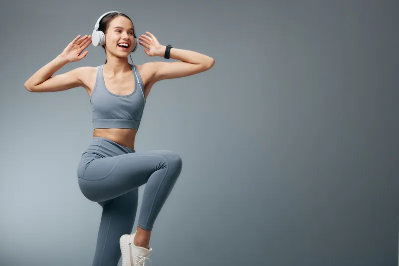 Woman fitness athlete in gray activewear with headphones, mid jump and raised knee against a neutral studio backdrop, capturing energy, movement, and a sporty training vibe.