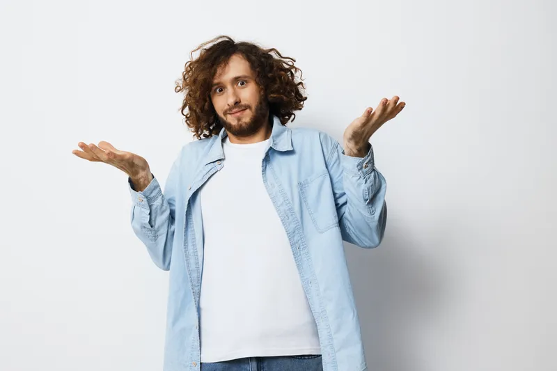 Young man with curly hair and a puzzled expression in a light blue shirt, conveying confusion and uncertainty against a minimalistic white background