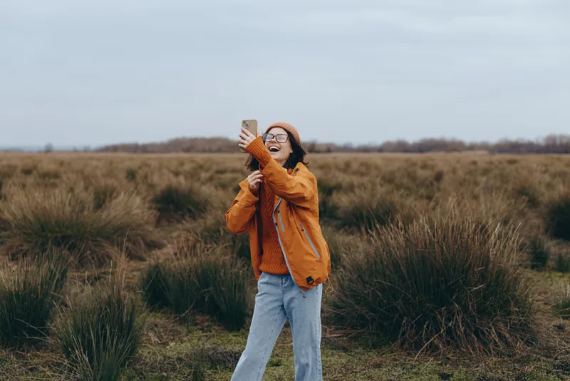Woman lifestyle outdoors, walking in grass field, orange jacket, smile on face while adjusting sunglasses, gentle overcast daylight, calm nature background, casual jeans