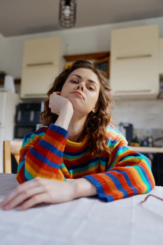 Woman in a colorful striped sweater sits at a kitchen table resting her chin on her hand, capturing a calm indoor lifestyle moment at home, candid and warm.