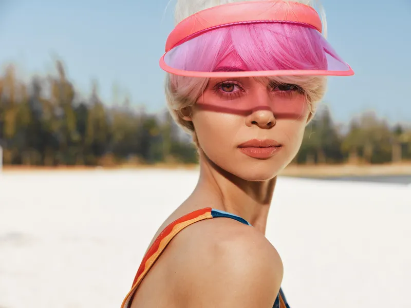 Portrait of a stylish woman on a sunny beach wearing a pink visor, visible swimsuit straps, and a soft breeze creating a relaxed, bright summer atmosphere.